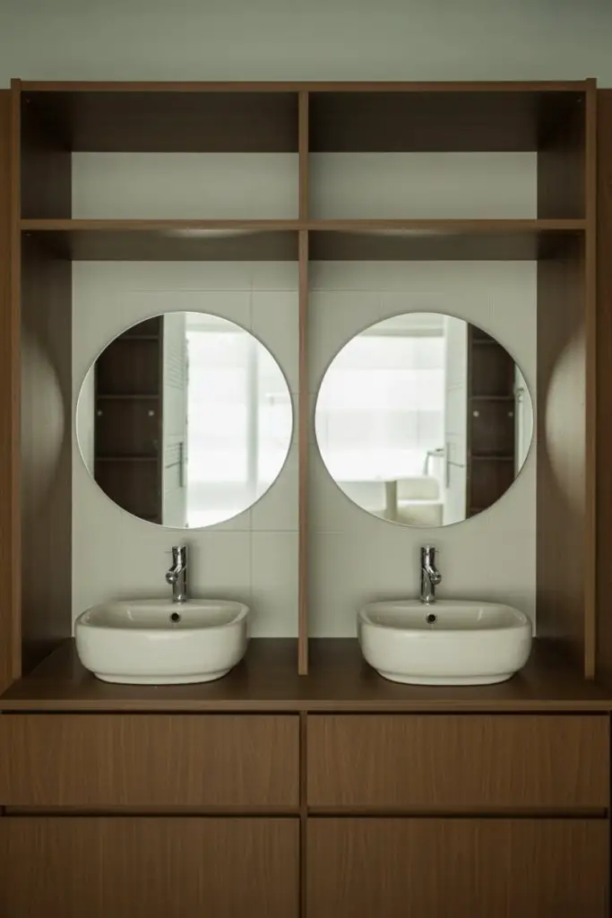 A modern bathroom with two round mirrors above two white sinks set in a wooden vanity, featuring open shelves and drawers beneath the sinks. The background shows a tiled wall reflected in the mirrors.