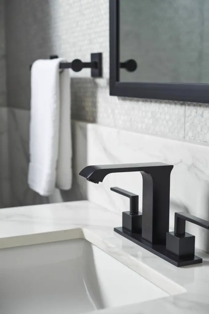 A modern bathroom with a black faucet on a white marble sink, a towel hanging on a black rod, and a large mirror with a black frame on a tiled wall.